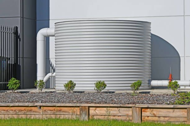 Large, corrugated metal water tank with white pipes against a building, sitting in a garden bed with small bushes.