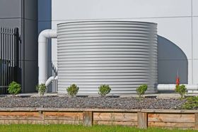 Large, corrugated metal water tank with white pipes against a building, sitting in a garden bed with small bushes.