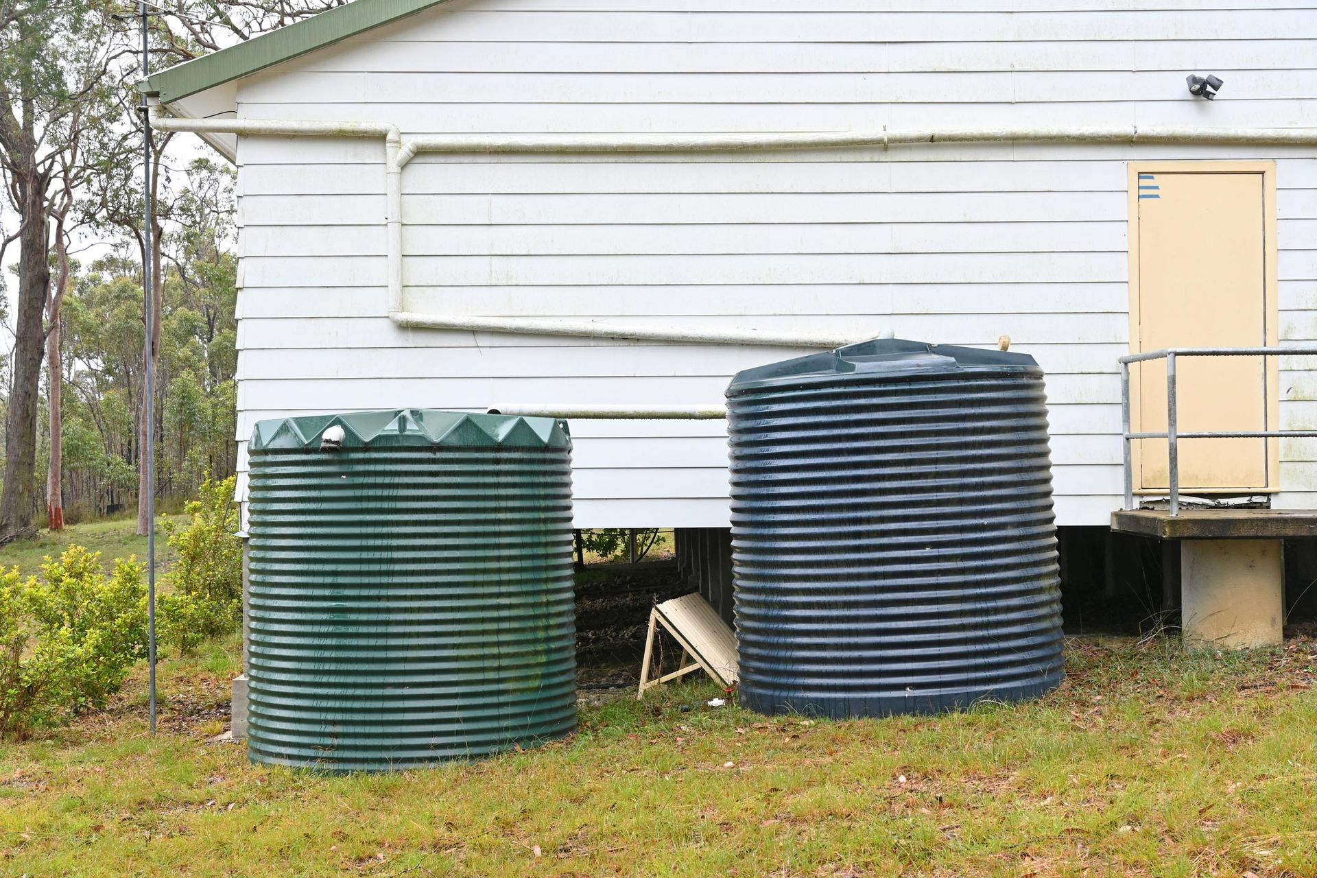Two corrugated metal water tanks, one green, one blue, next to a white house with a beige door.