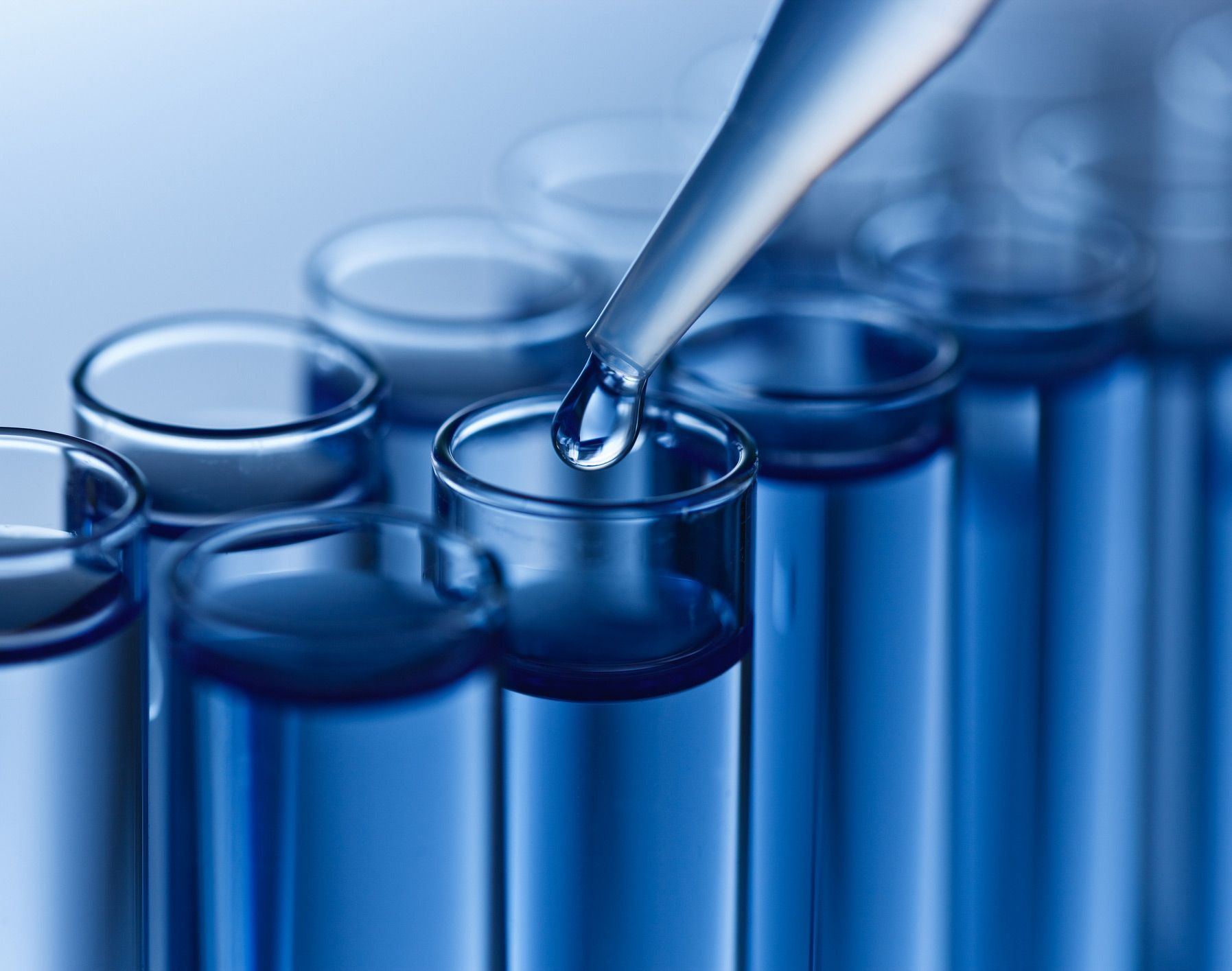 A dropper dispensing liquid into a test tube with other test tubes in the background, all filled with blue liquid.