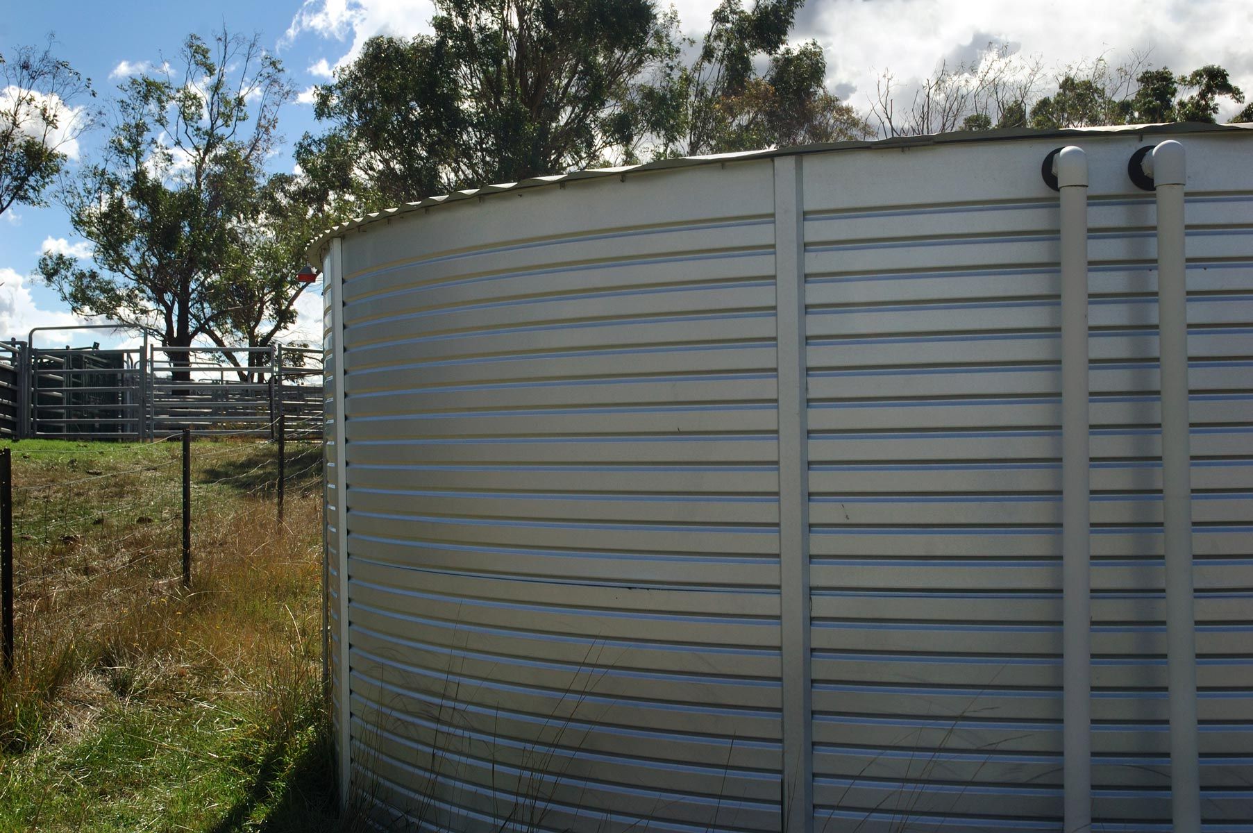 Large, cylindrical metal water tank outdoors on a grassy field; trees and a fence in the background.