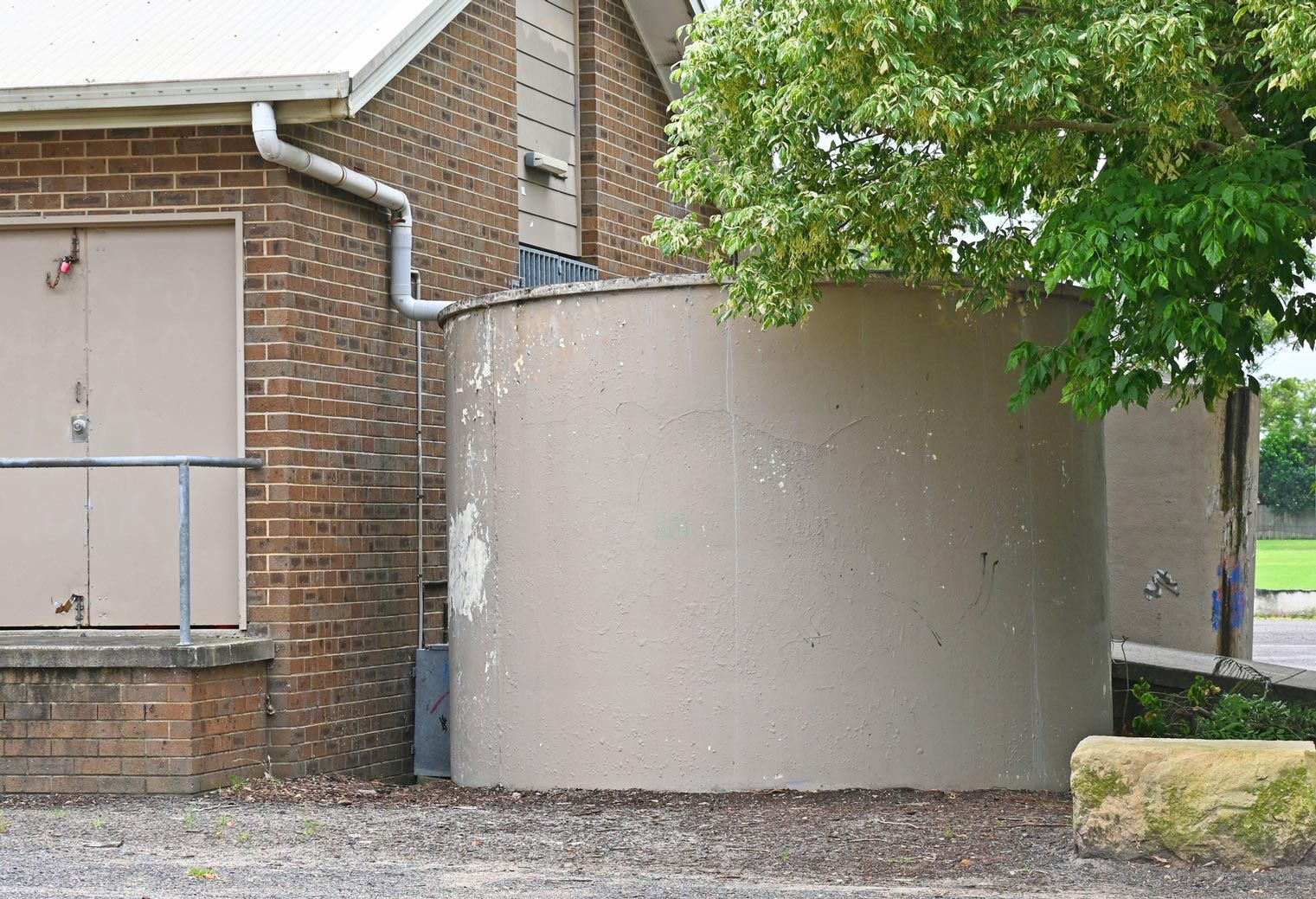 A tan cylindrical water tank next to a brick building, with a downspout and tree visible.