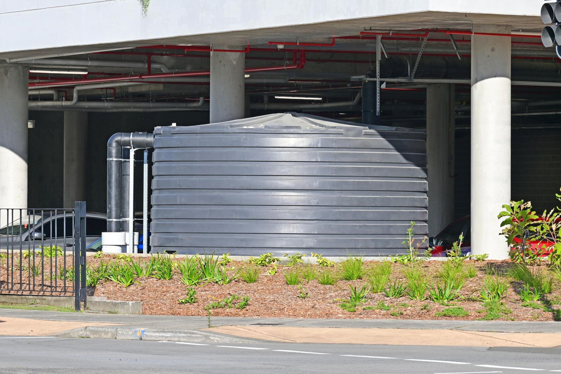 Black water tank under a building with pipes, surrounded by greenery and concrete.