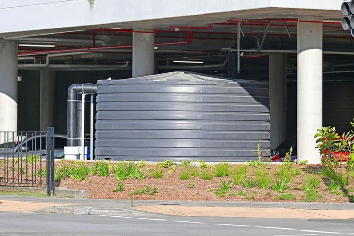 Black water tank under a building with pipes, surrounded by greenery and concrete. Black water tank under a building with pipes, surrounded by greenery and concrete.