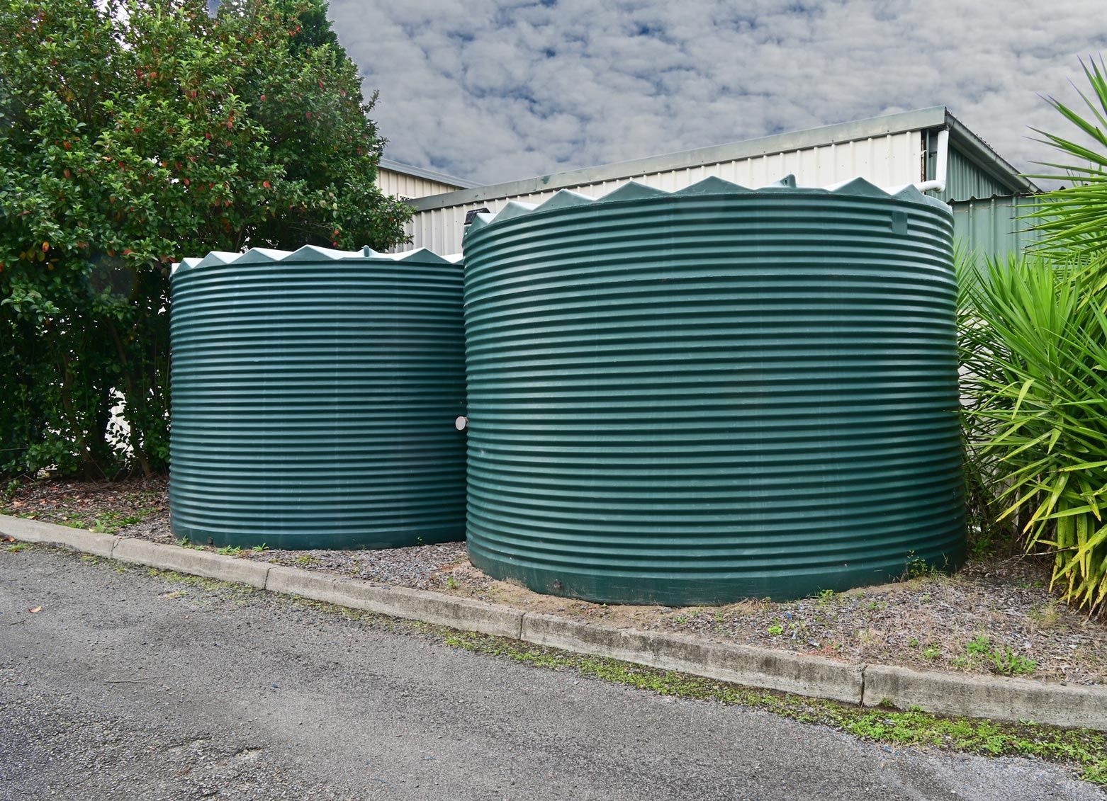 Two green corrugated metal water tanks on a concrete pad, surrounded by greenery and a building in the background.