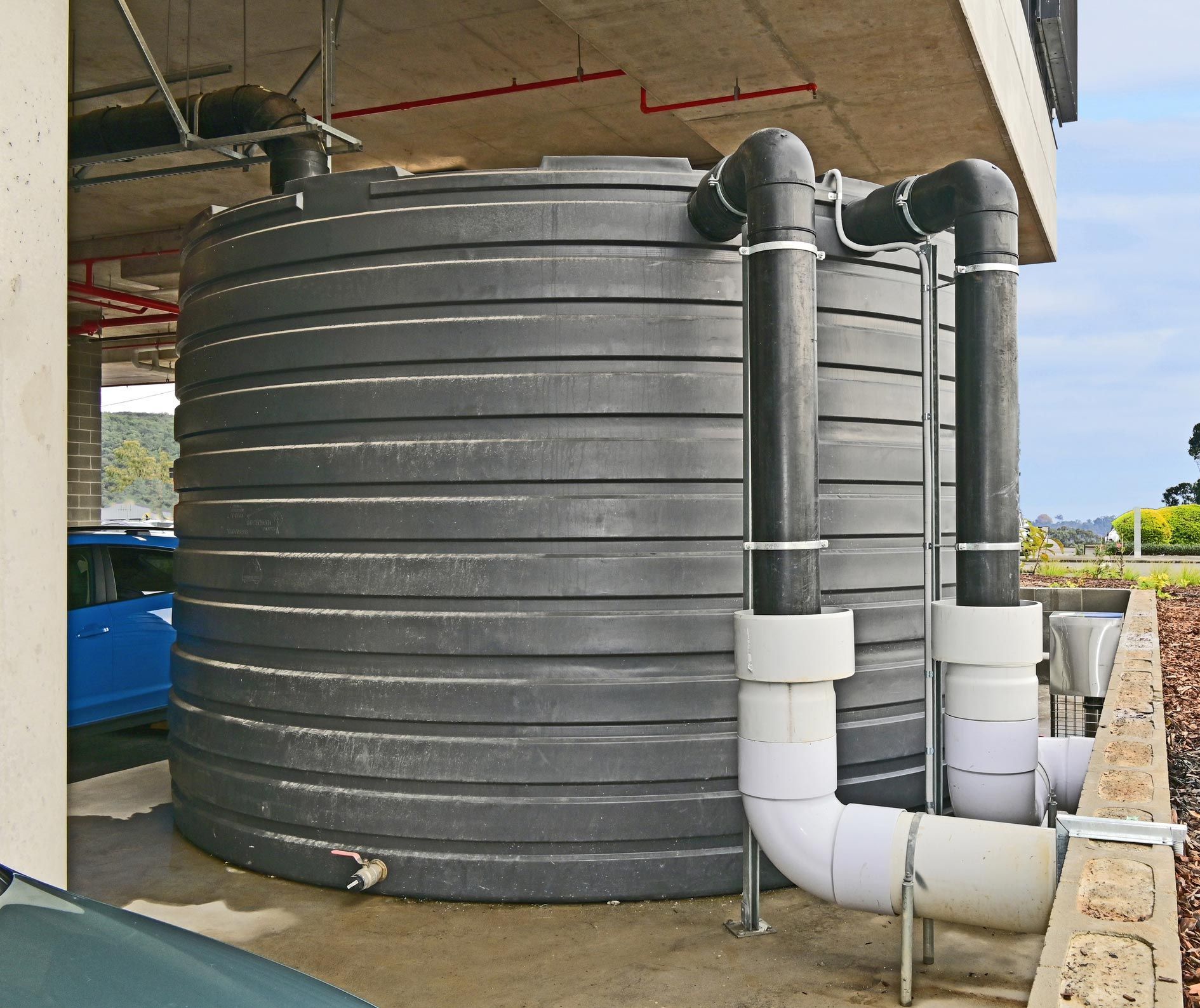 Large, black water storage tank with pipes, outdoors under a concrete structure.