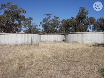 Three concrete water tanks in a grassy field, trees in the background, blue sky overhead.