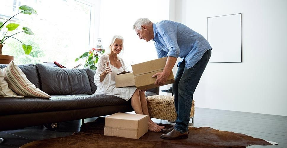 A man is lifting a box in a living room while a woman sits on a couch.