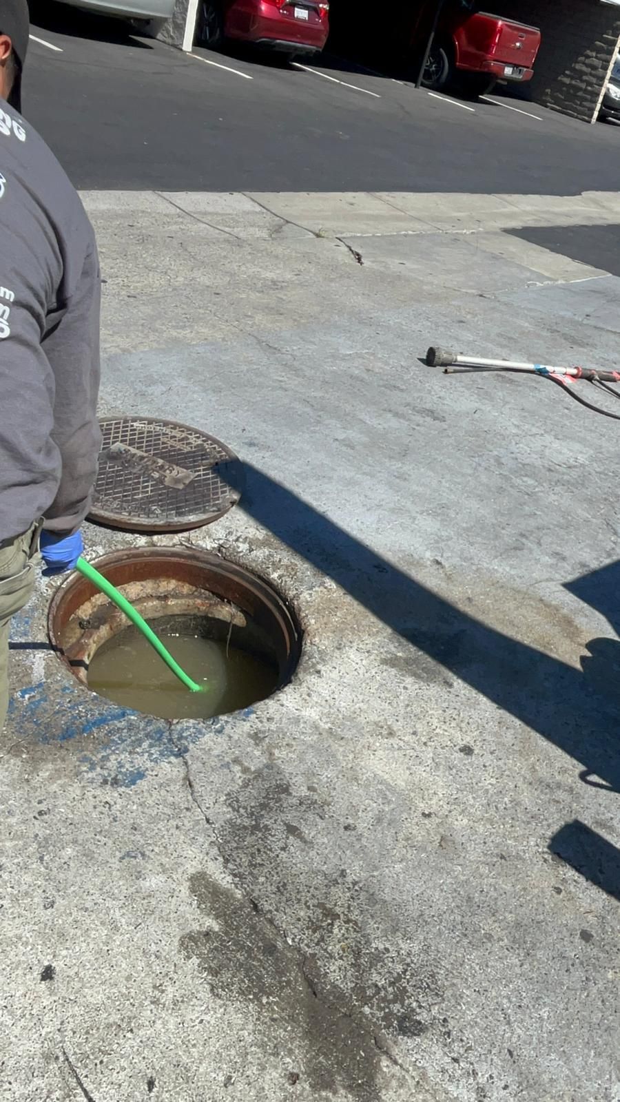A person is working on an open sewer drain in the street with water inside, gray concrete and cars in background.