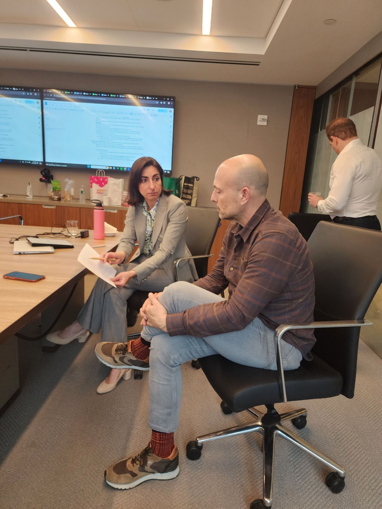 Man in a brown blazer, blue shirt, speaking with hands raised, on a stage with a dark curtain.