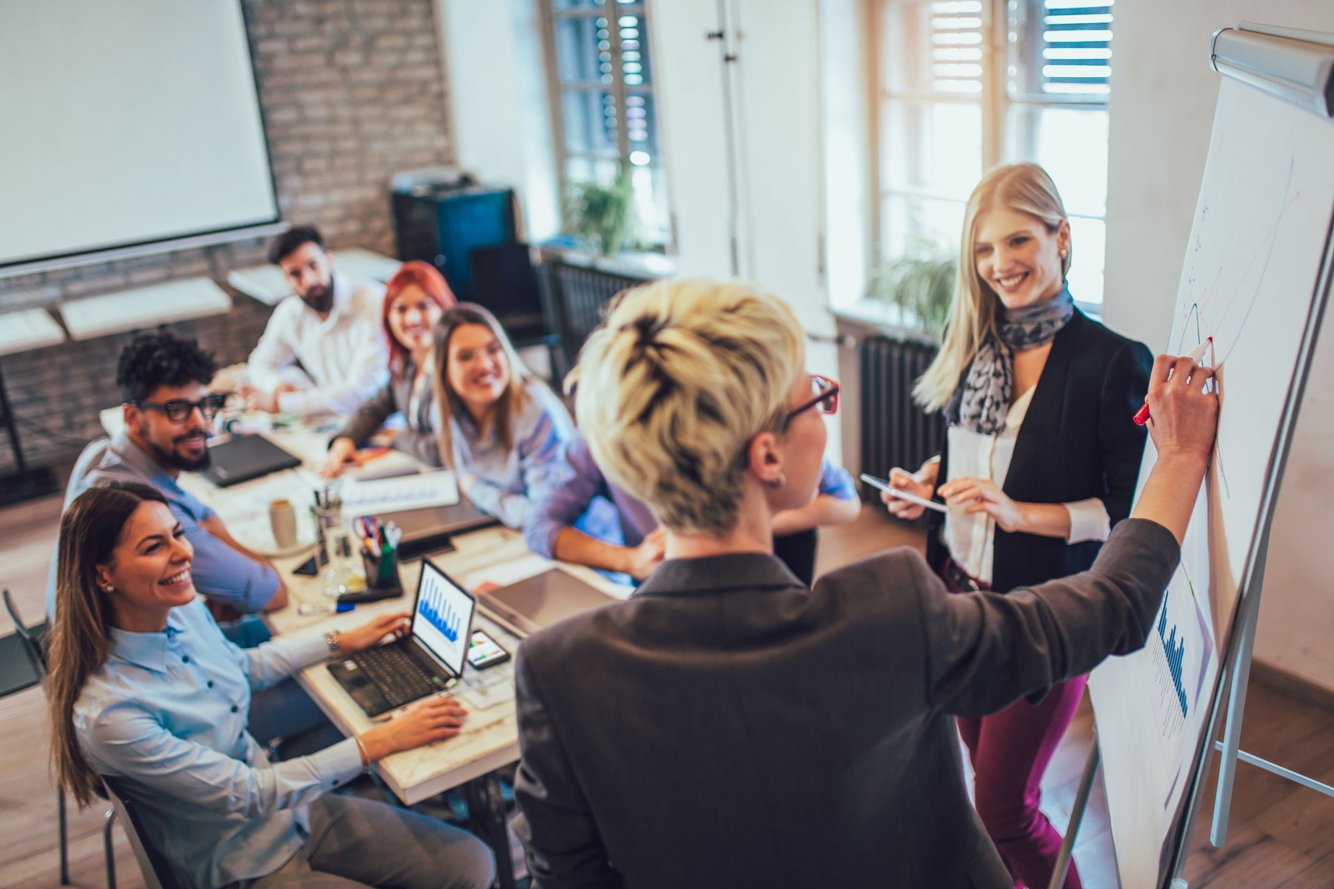 Woman presenting at a whiteboard to a group in an office. They are seated around a table, smiling. Woman presenting at a whiteboard to a group in an office. They are seated around a table, smiling.