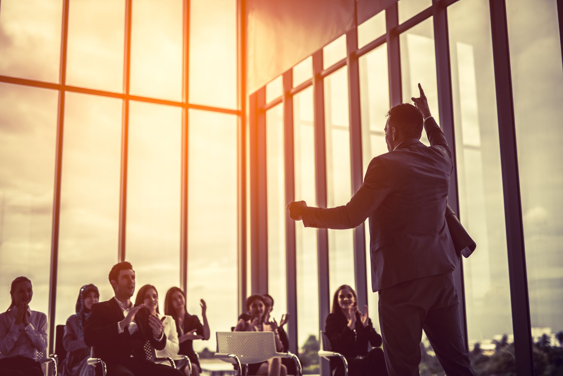 Man in suit giving presentation, gesturing towards large windows, with audience clapping.