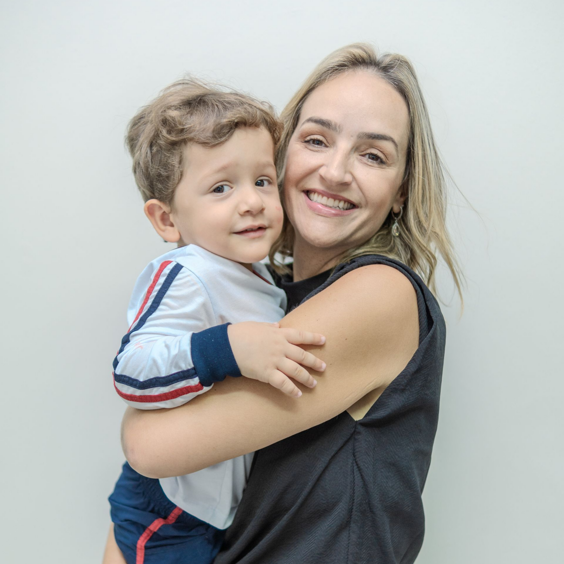 Mulher segurando uma criança sorridente; ambas olham para a câmera. Parede branca ao fundo.