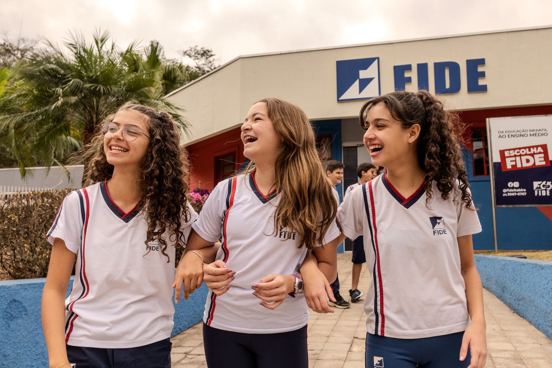 Três estudantes com uniformes escolares brancos e azuis riem e caminham juntos em frente a um prédio com uma placa da FIDE.