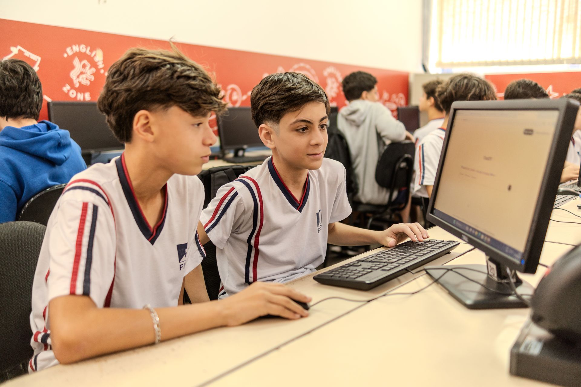 Dois meninos em uniformes escolares trabalhando em um computador em uma sala de aula.