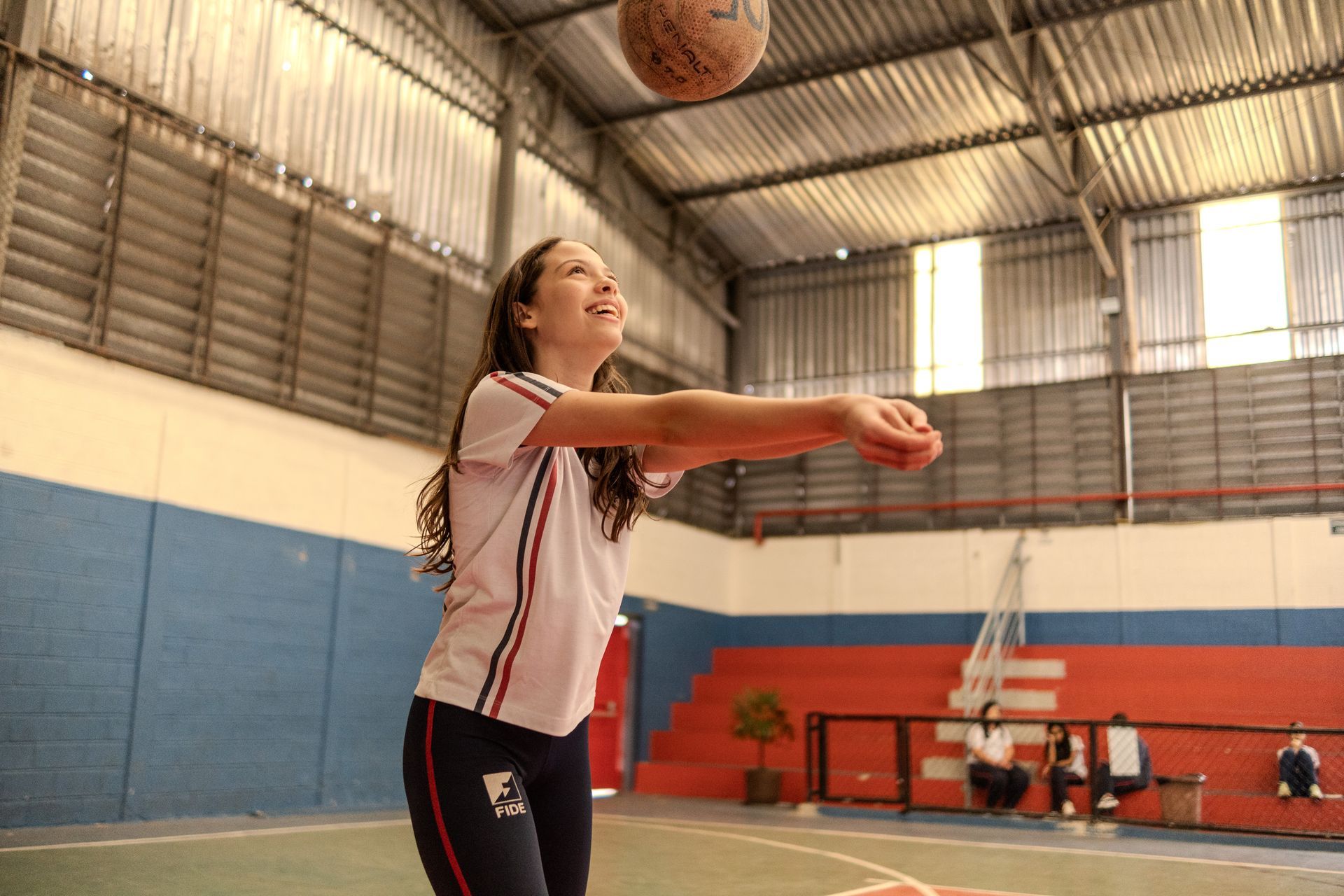 Uma jovem mulher de uniforme escolar jogando vôlei, sorrindo, em uma academia coberta.