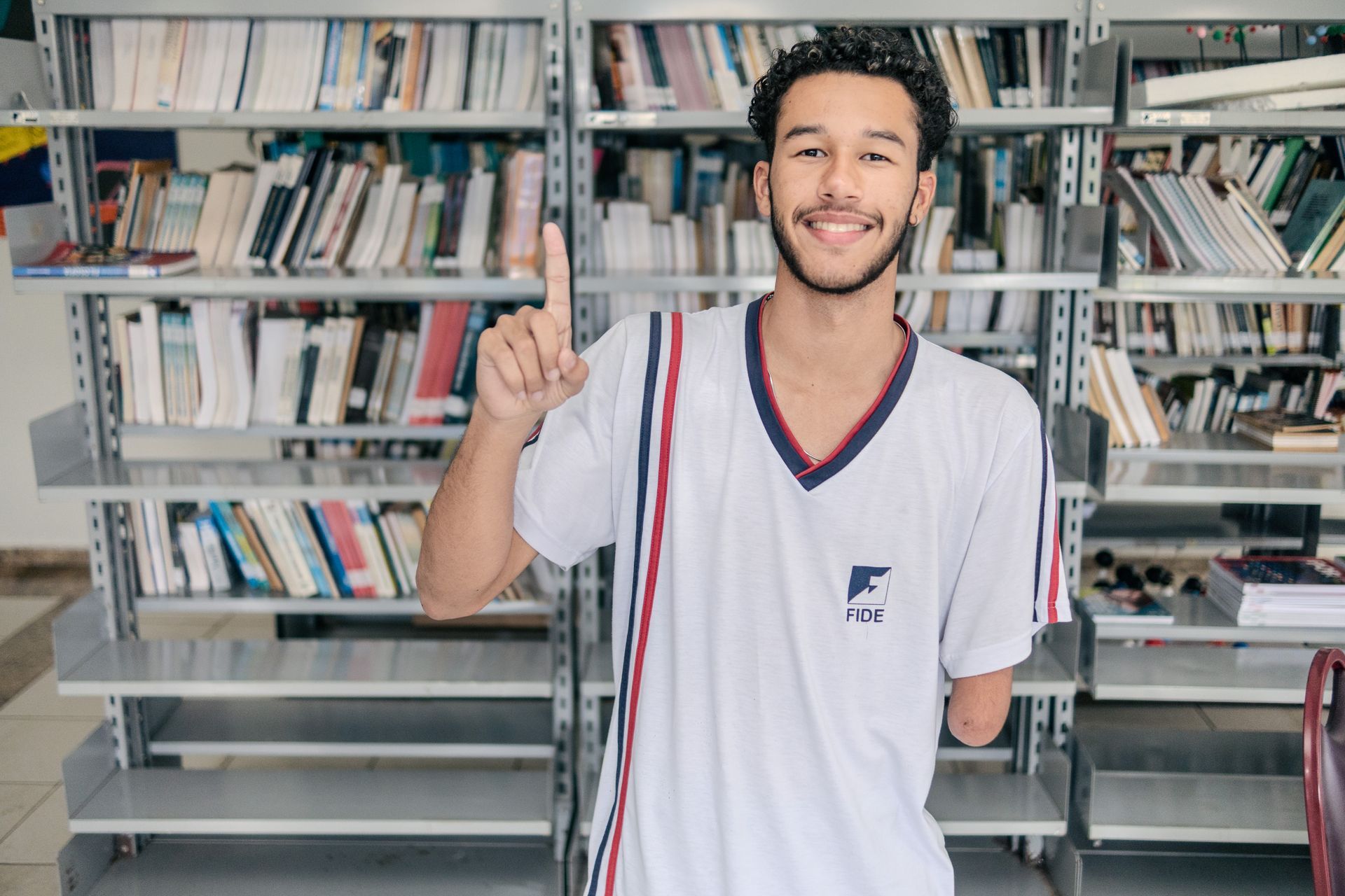 Jovem sorridente com um braço apontando para cima em uma biblioteca, vestindo uniforme escolar.