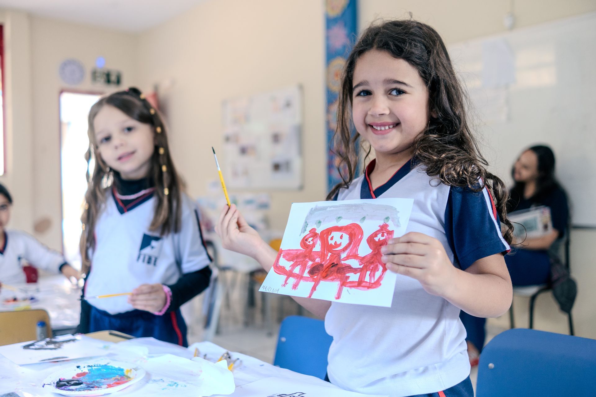 Duas meninas sorridentes em uma aula de arte, uma delas segurando uma pintura com tinta vermelha; ambiente de sala de aula.