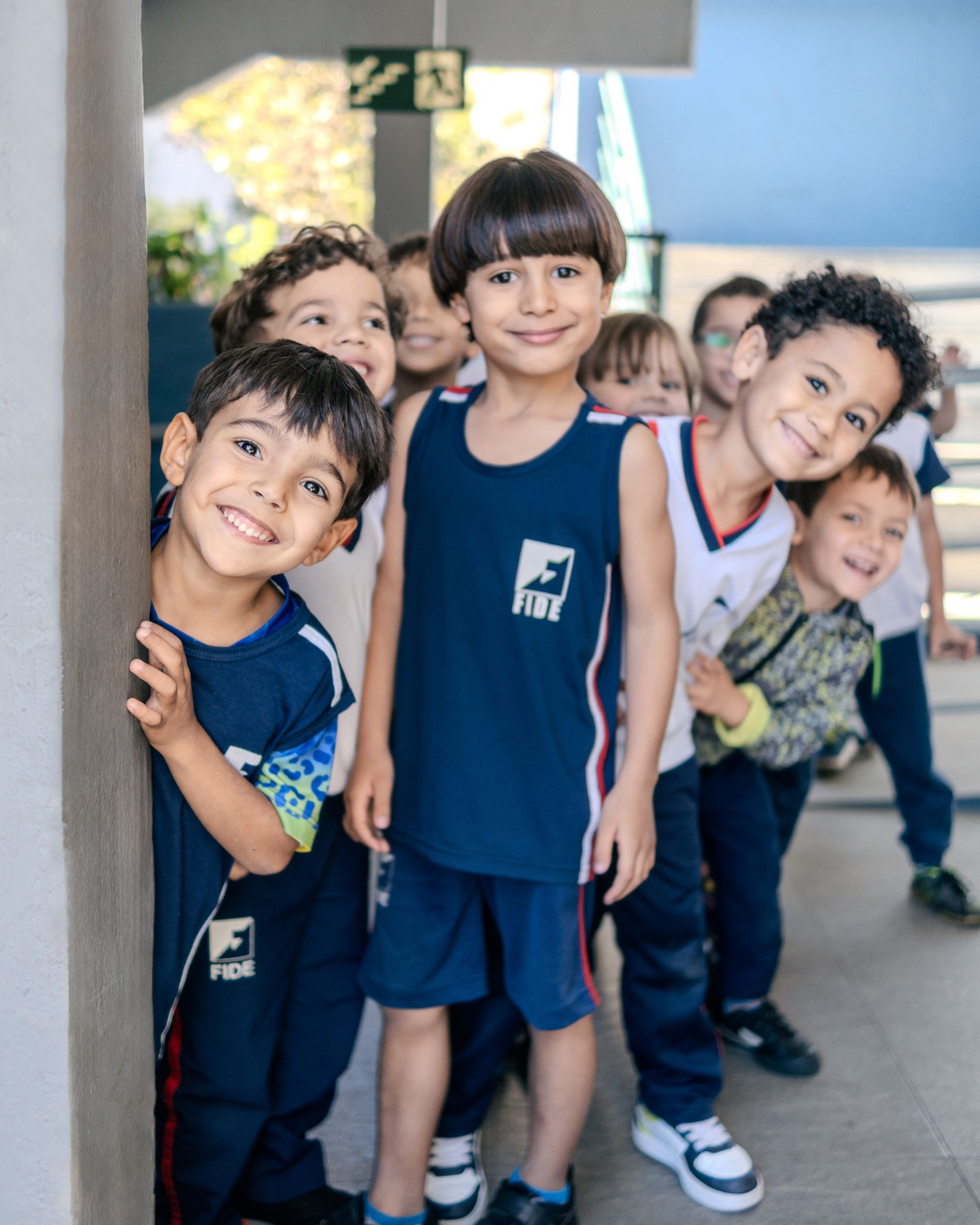 Crianças em uniformes escolares sorriem e espiam por trás de uma parede em um corredor bem iluminado.