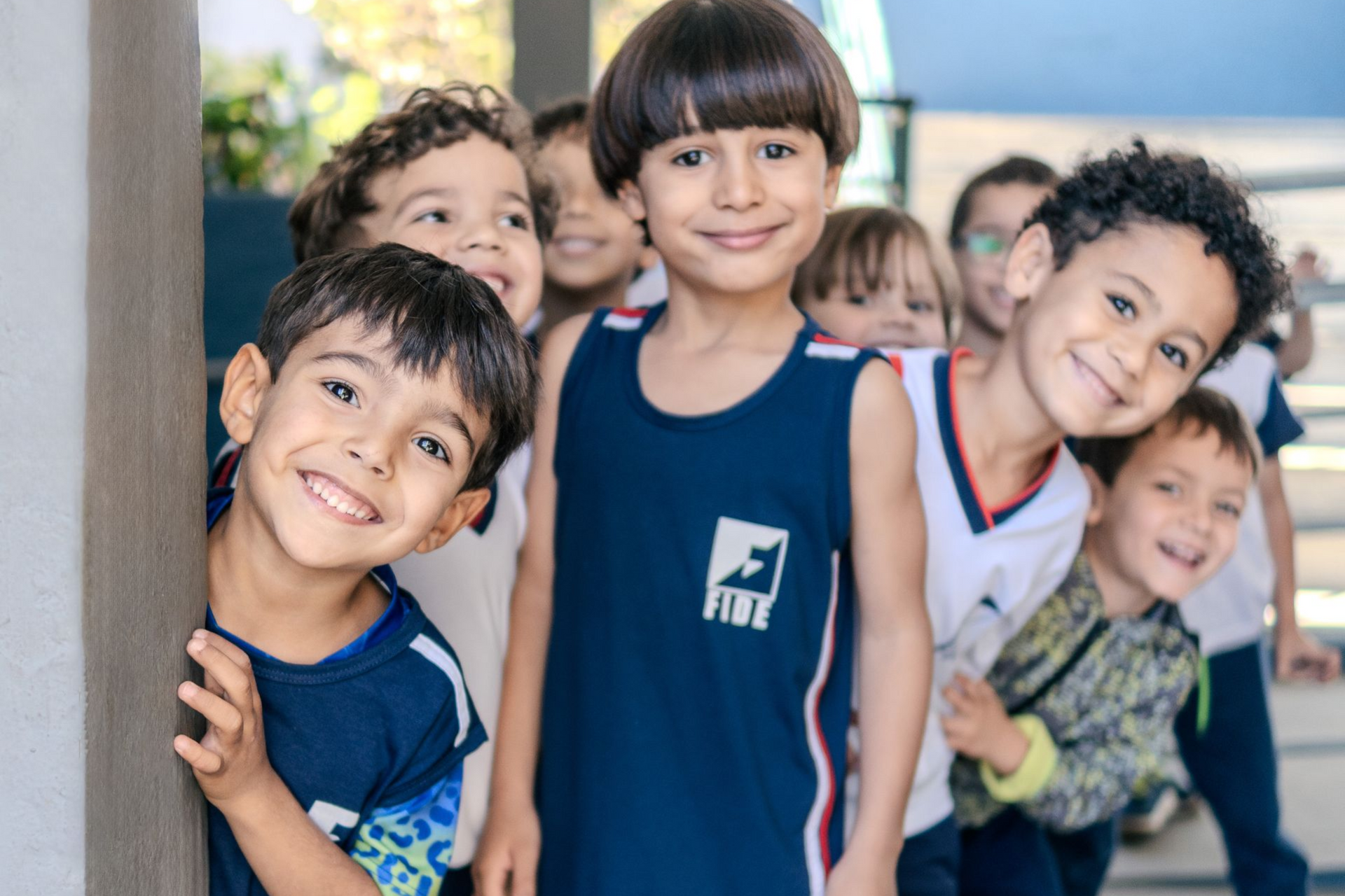Um grupo de estudantes sorridentes, trajando uniformes escolares, está em fila enquanto espia por trás de um pilar.
