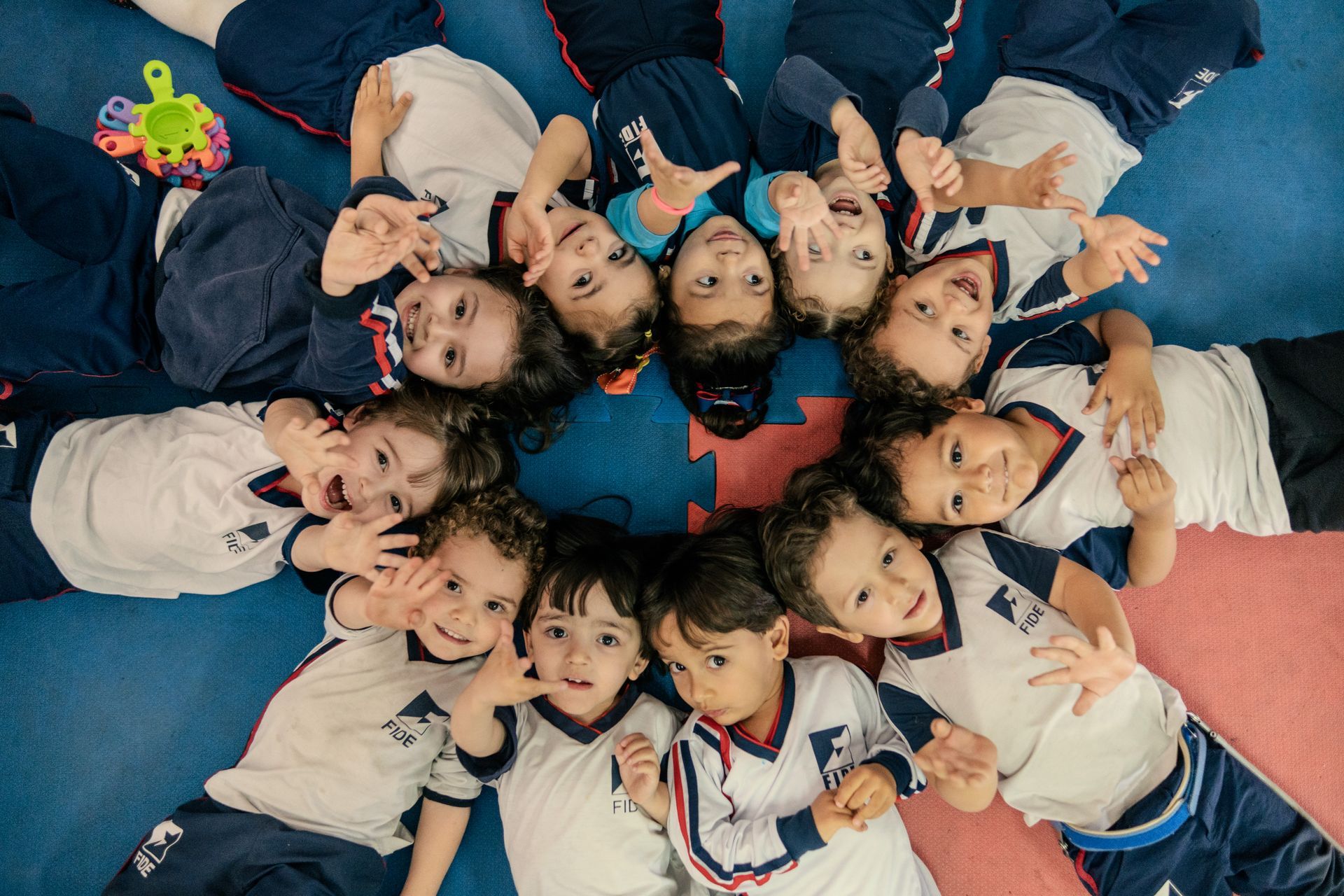 Um grupo de crianças com uniformes brancos e azuis está deitado em círculo sobre um tapete azul, sorrindo e acenando para a câmera.