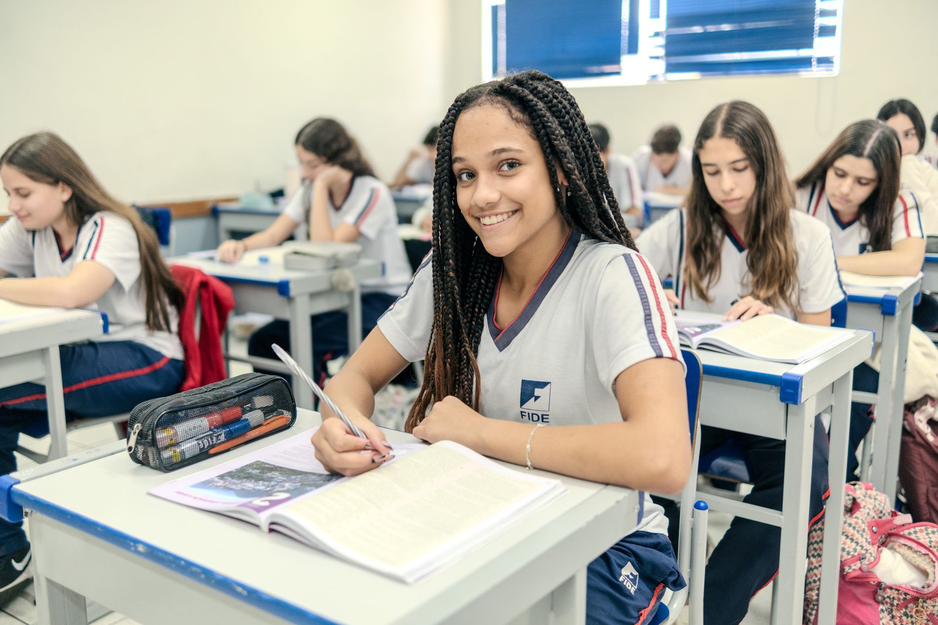 Um aluno sorrindo em sua carteira em uma sala de aula bem iluminada, escrevendo em um caderno aberto entre colegas uniformizados.