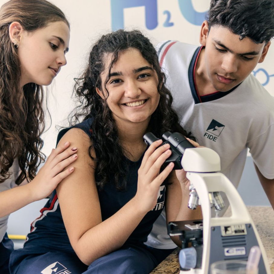 Três estudantes olhando para um microscópio em um laboratório, um sorrindo, os outros olhando para a lâmina.