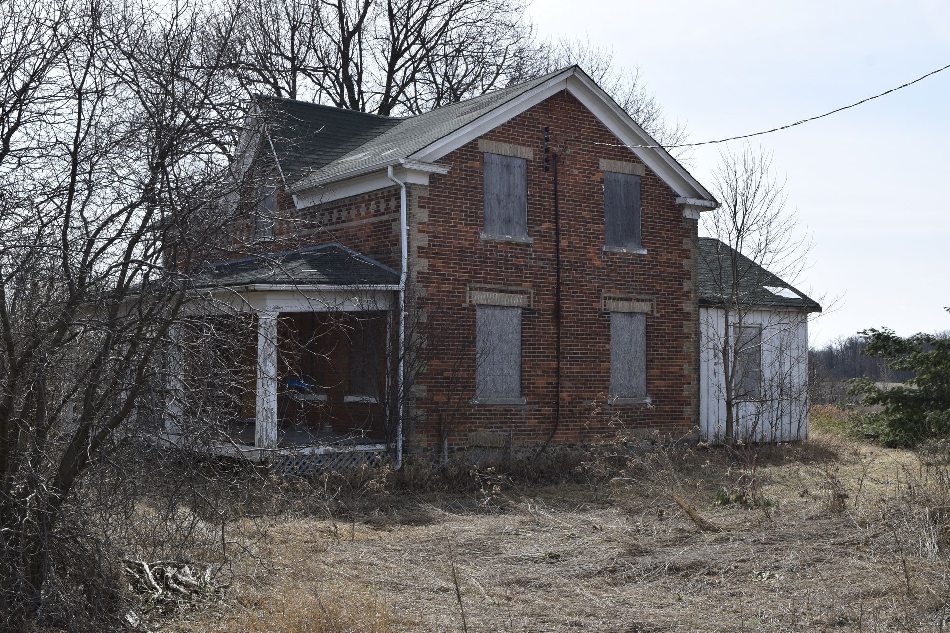 Abandoned brick gothic revival farmhouse with boarded windows in an overgrown yard 