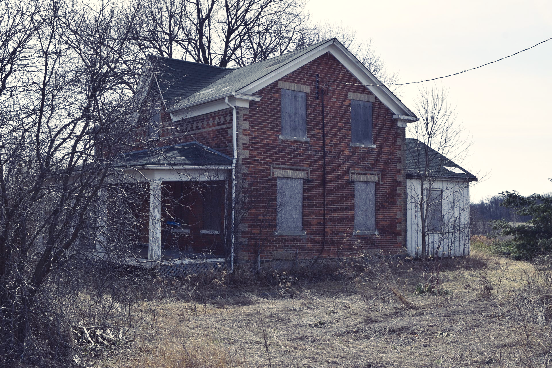 Abandoned gothic revival brick farmhouse with boarded windows in an overgrown yard