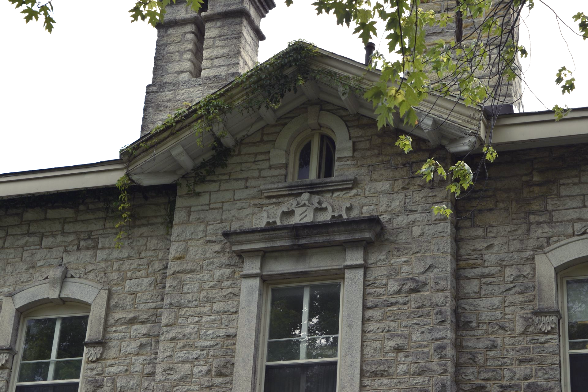 Stone building facade with arched windows, decorative trim, and a central dormer under leafy branches