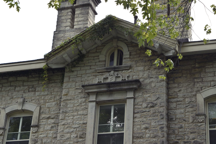 Stone building facade with arched windows, decorative trim, and a central dormer under leafy branches