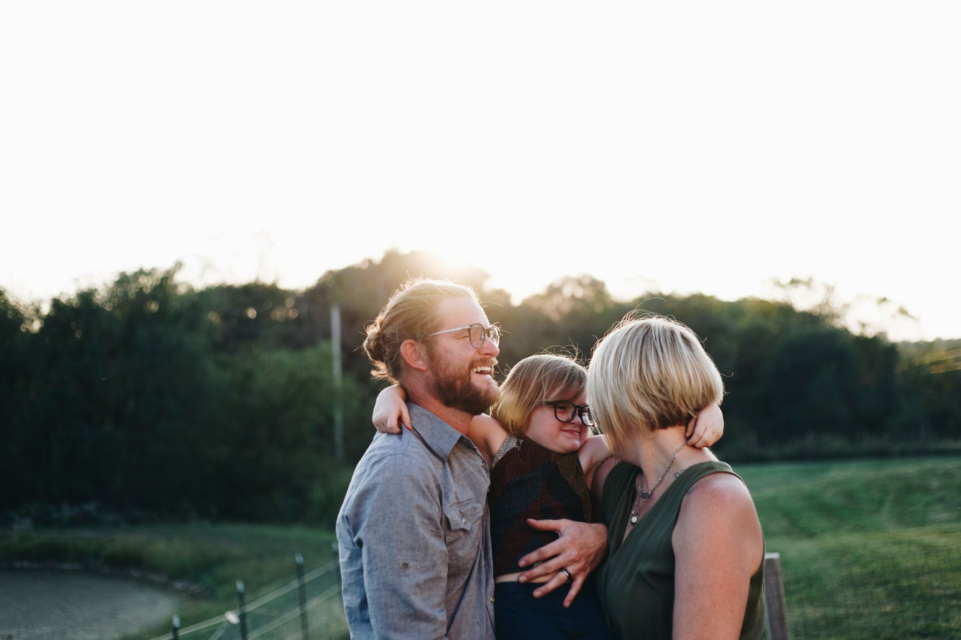 Family of three embracing outdoors, smiling, backlit by sun.