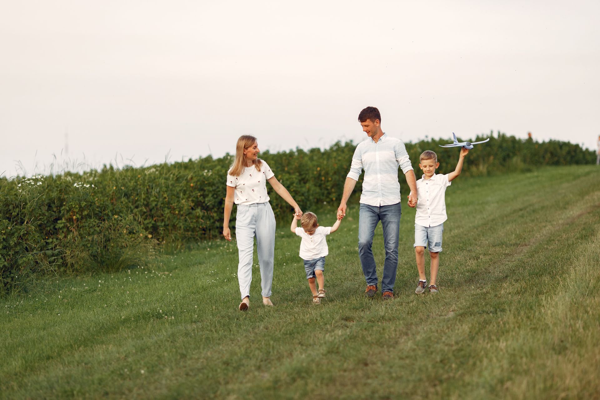 Family walking on a grassy hill, holding hands. A child holds a toy airplane. Overcast sky.