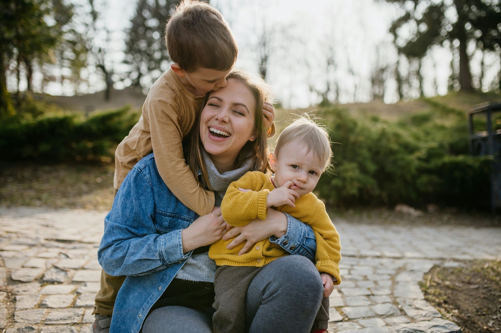Woman laughing with two children in a park; one kisses her head, the other cuddles, smiling.