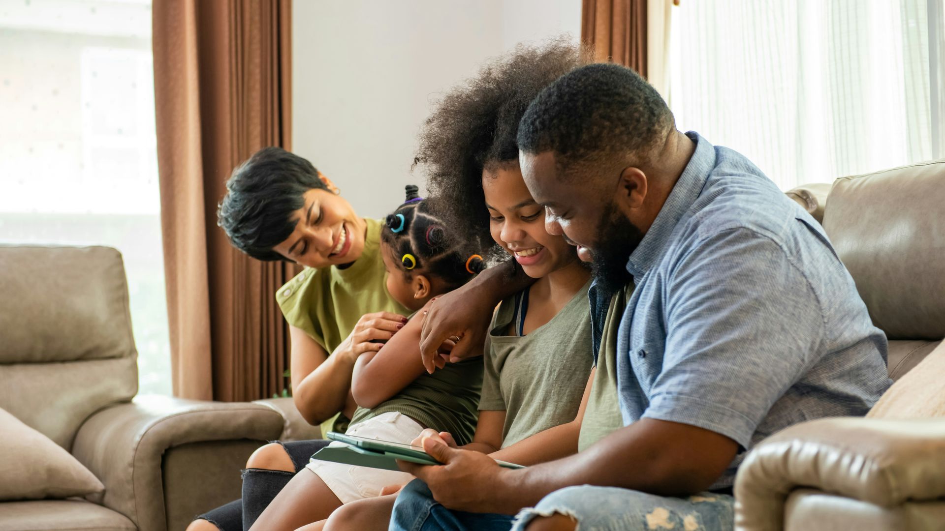 Family of four sitting on a couch, looking at a tablet together, smiling. Living room setting.
