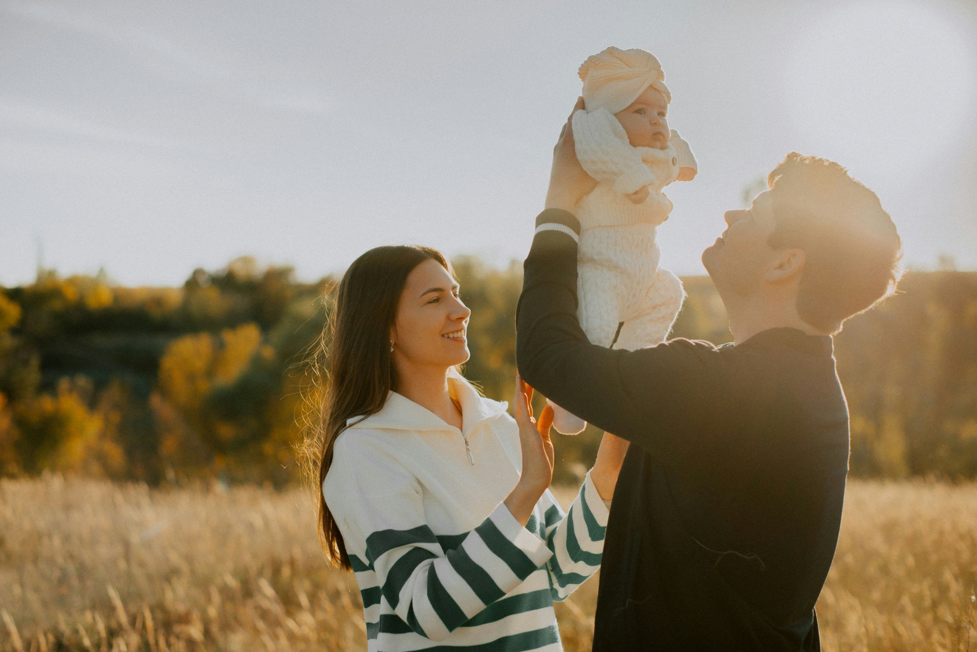 Parents holding a baby up in the air in a field. Golden light and greenery in background.