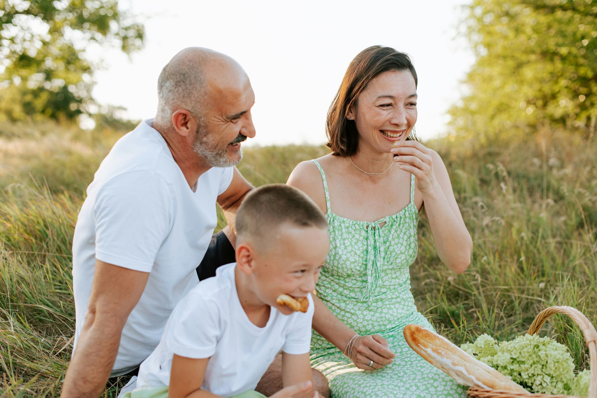 Family on a picnic, laughing and eating outdoors. A basket with food is nearby.