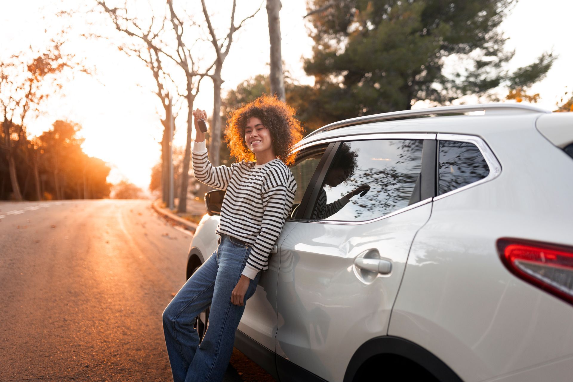 Woman leaning on white car on road, smiling, holding keys; sunset.