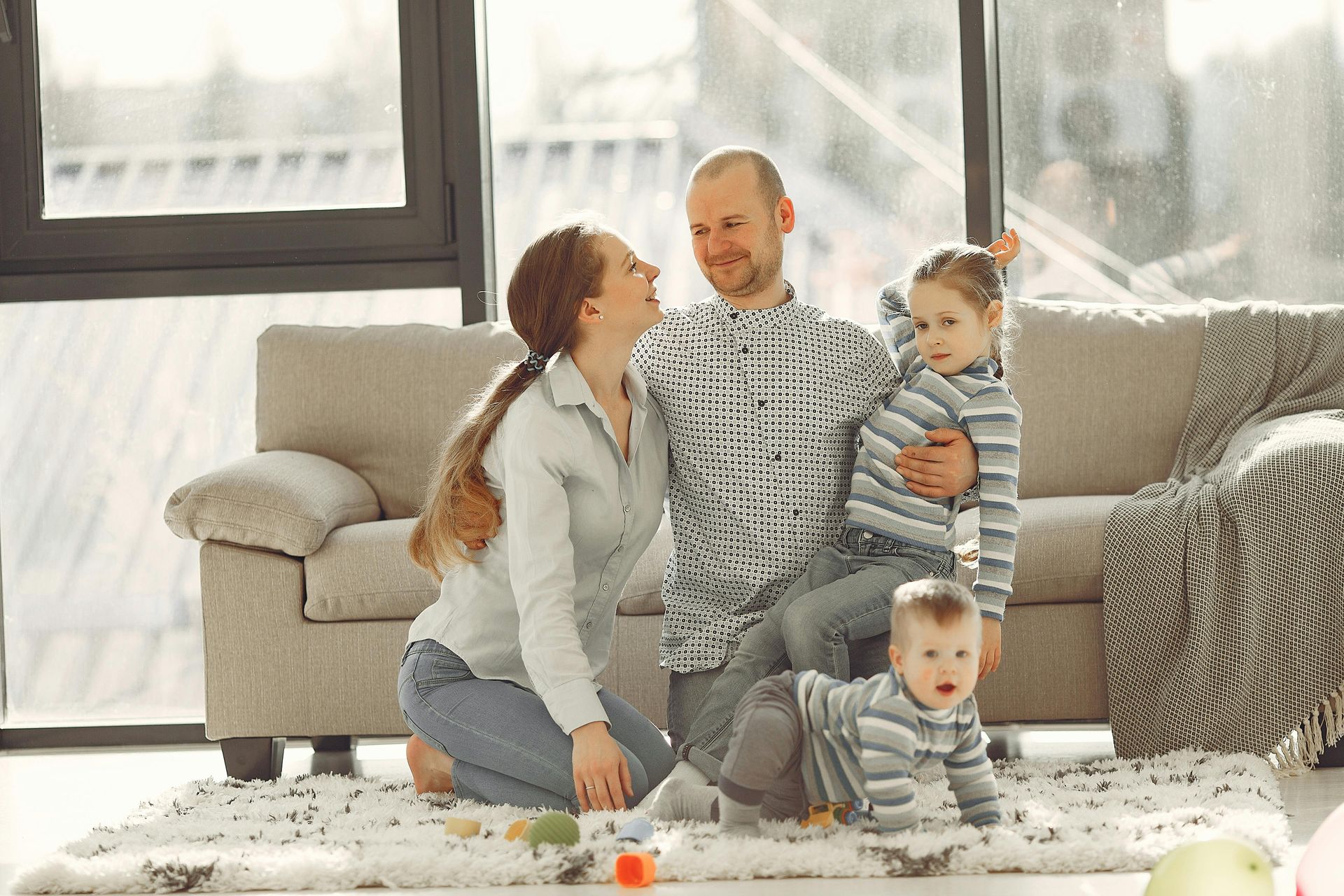 Family of four in living room, smiling near a sofa and window.