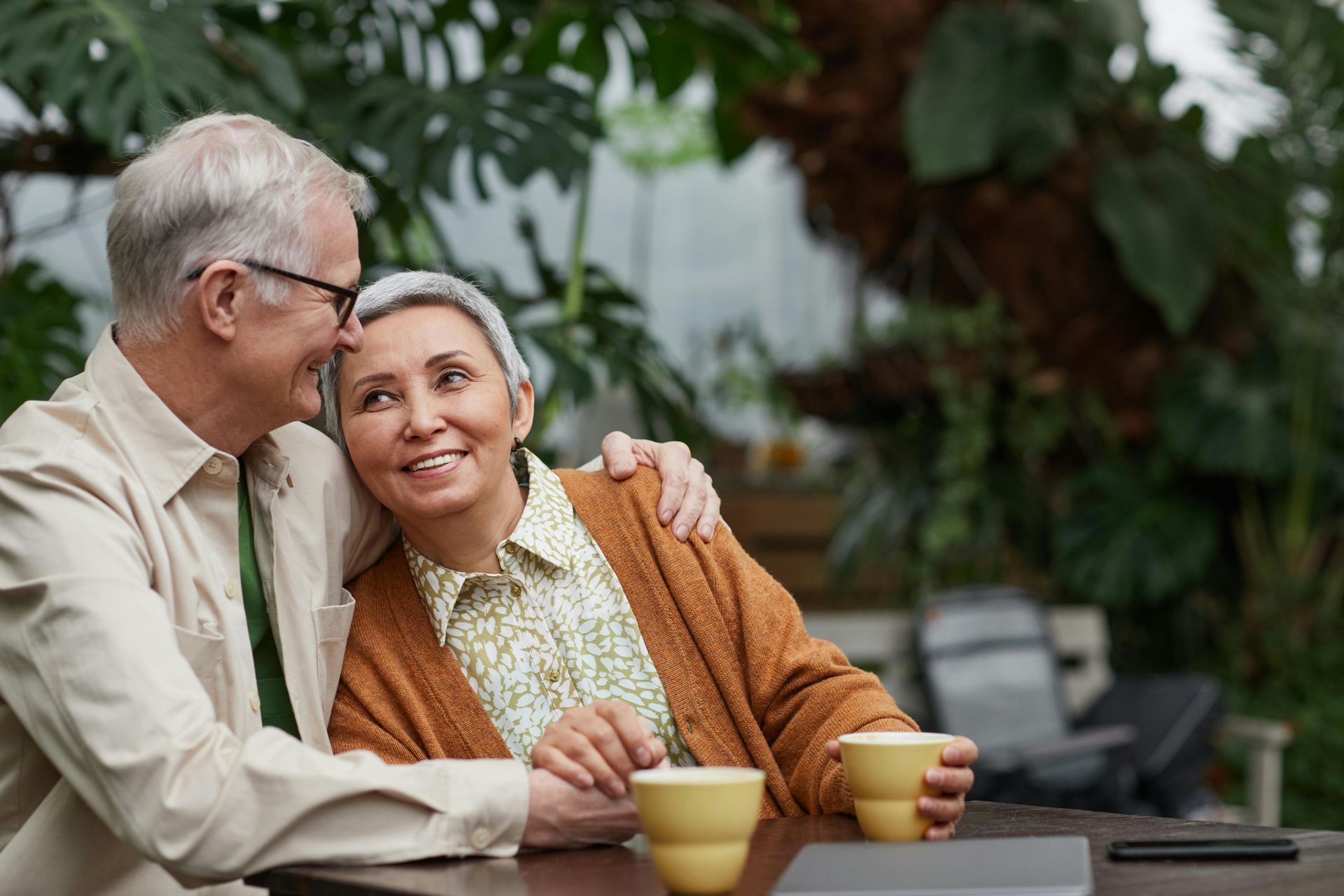 Elderly couple smiles, man's arm around woman's shoulders, sitting at a table with coffee cups, surrounded by plants.