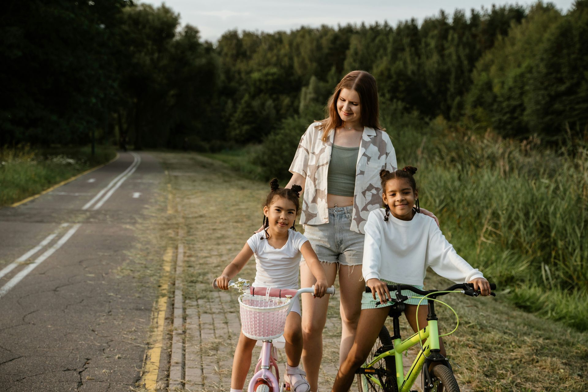 Woman with two children and bicycles on a paved path near trees.