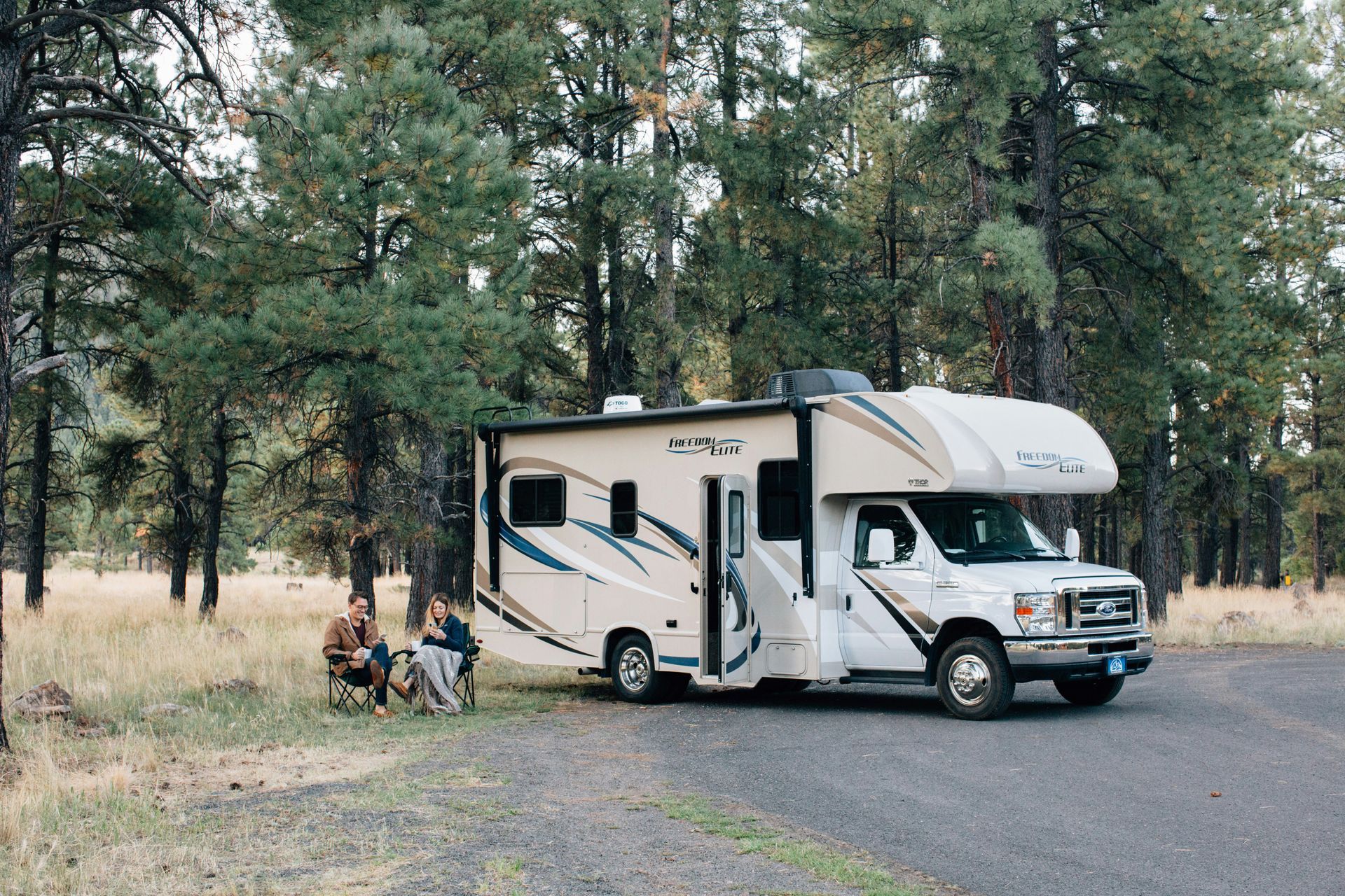 RV parked on gravel road near trees; people sitting in chairs nearby.