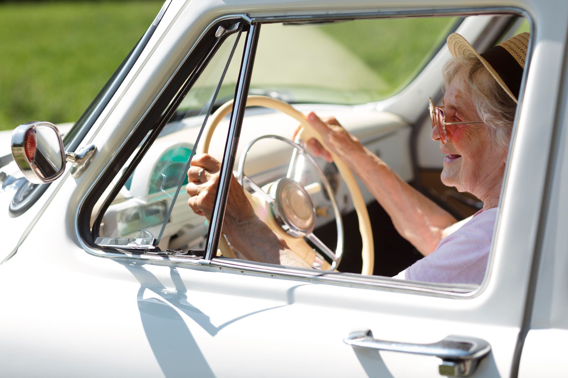 Elderly person in a white car, smiling, holding steering wheel. Wearing a hat and glasses.