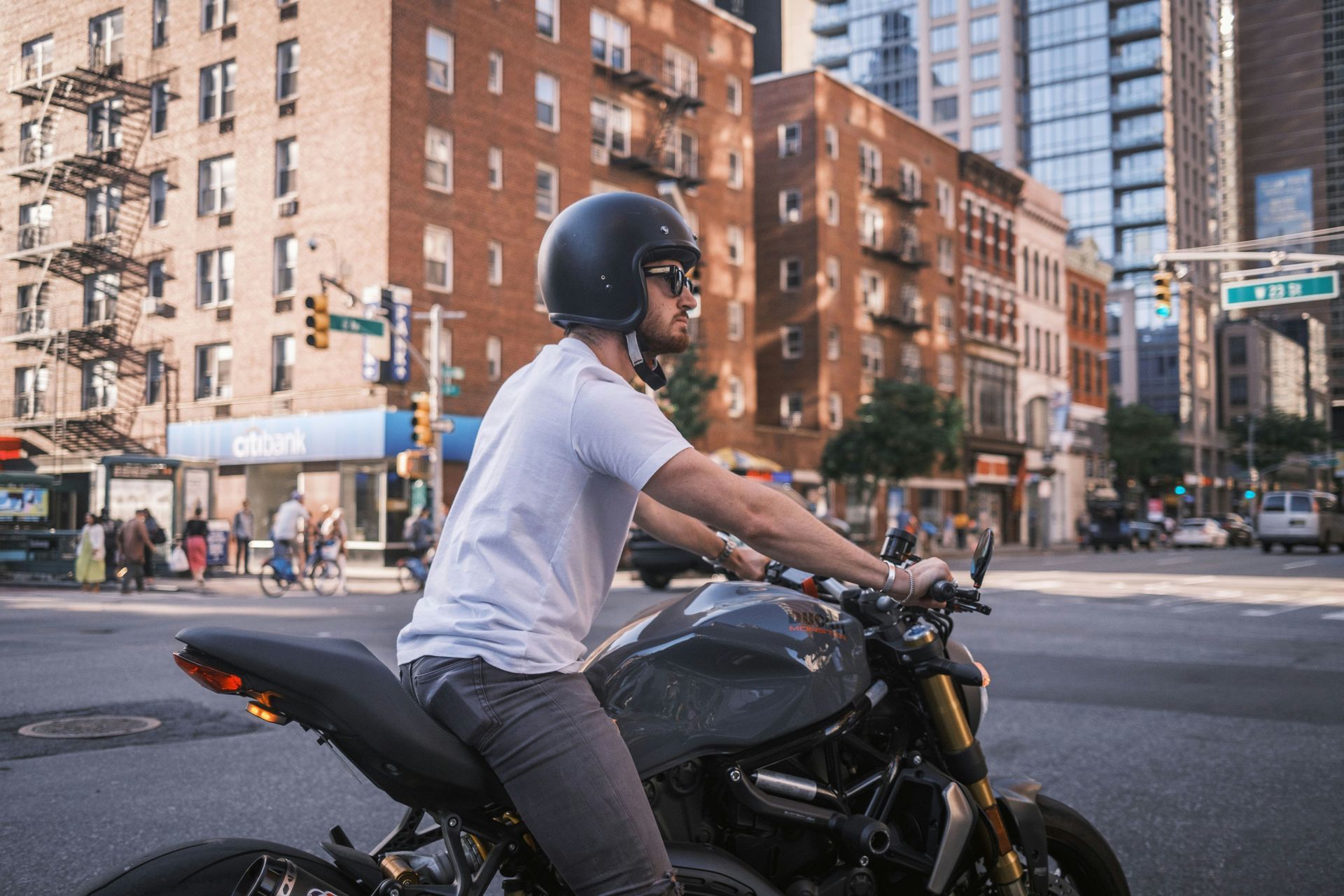 Man on motorcycle, wearing a helmet, stopped at a city intersection with brick buildings in the background.