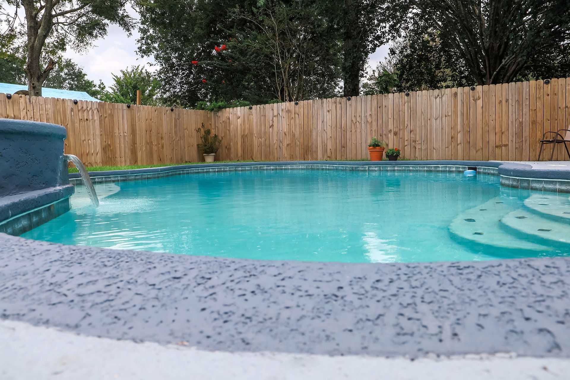 Backyard swimming pool with turquoise water, wooden fence, and trees under an overcast sky
