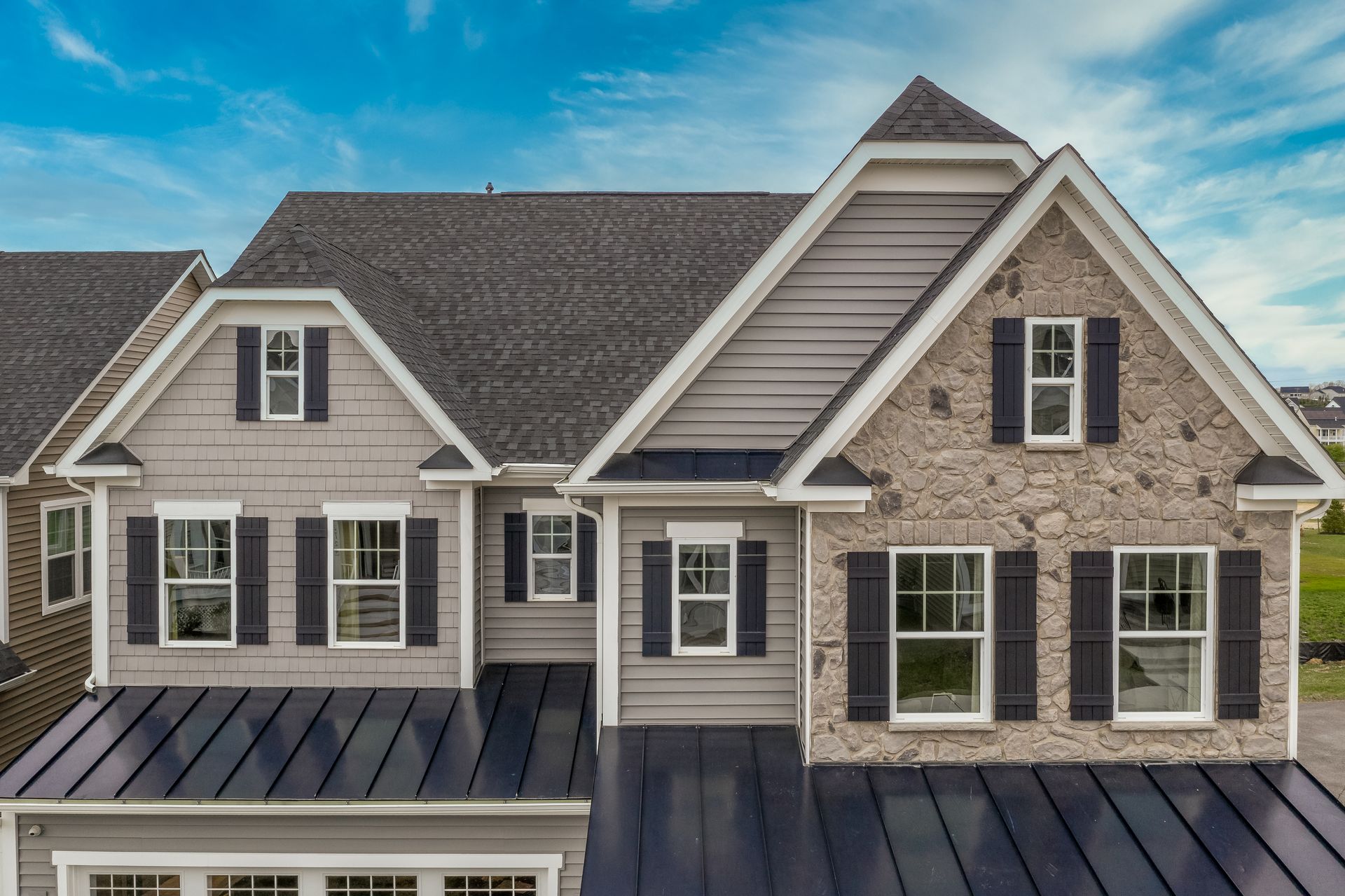 Suburban house exterior with gray siding, stone accents, and black metal roof under a blue sky