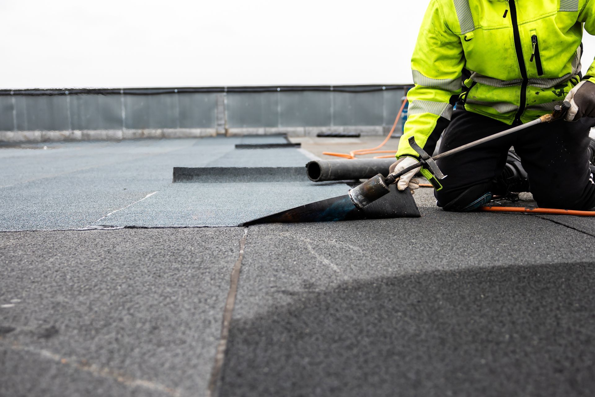 Worker installing roofing membrane on a flat roof with a torch, wearing a bright yellow safety jacket.