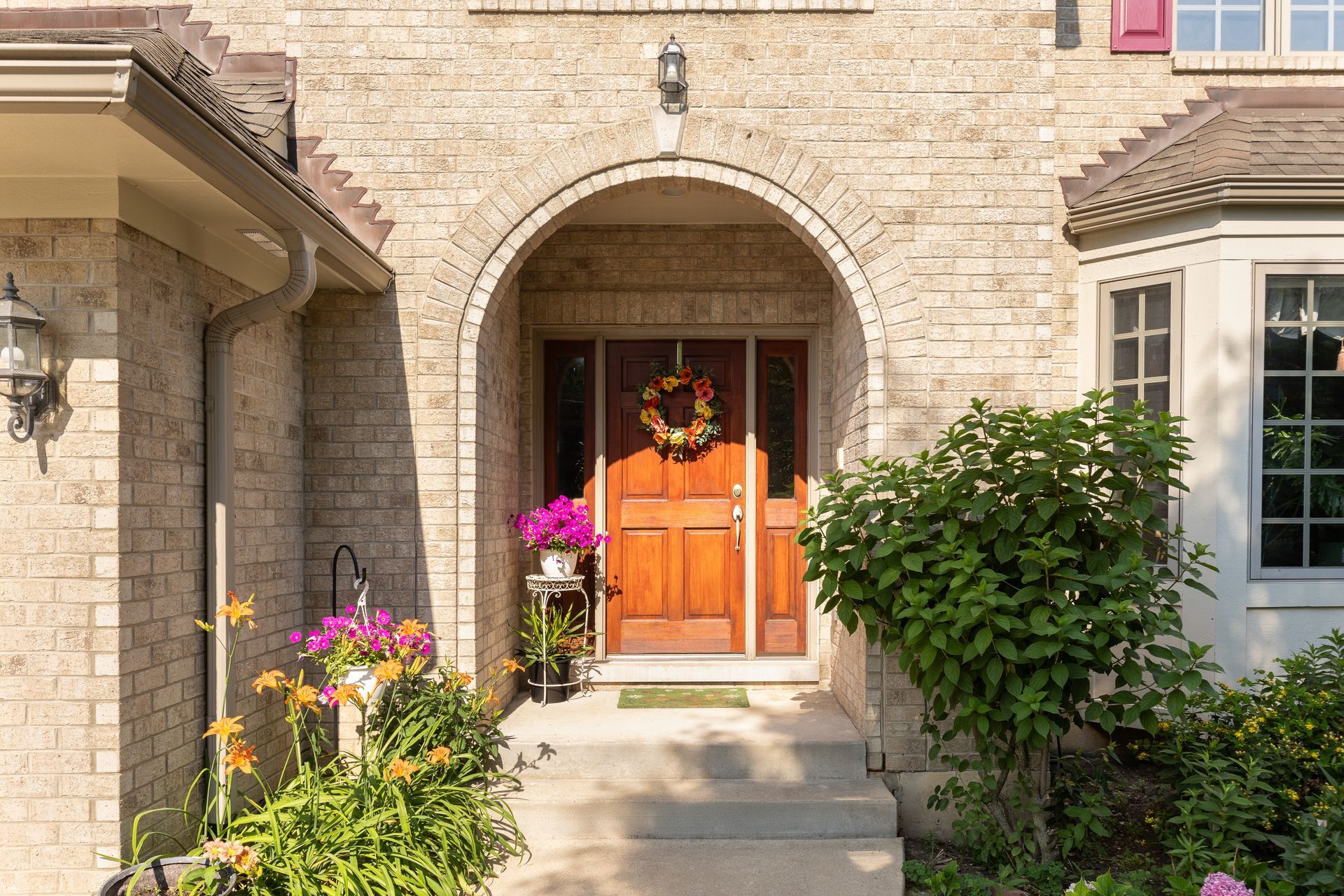 Brick house entryway with arched doorway, wooden double doors, and colorful flowers by the path