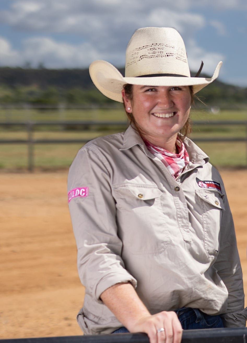 Woman in cowboy hat, smiling, outdoors, tan shirt, leaning on fence, dirt arena background.