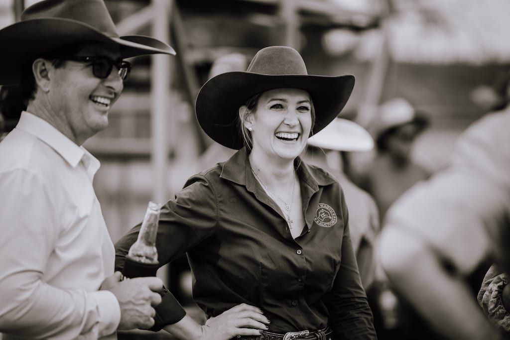 Two smiling people in cowboy hats, an outdoor event. Man holding a drink, woman laughing with hand on hip.